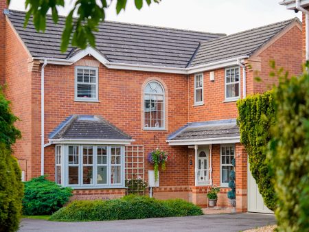Modern red brick house with white-framed windows, a small porch entrance, and a landscaped front garden with shrubs and hanging flowers.