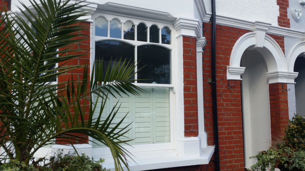 White framed bay window with decorative panes and shutters set in a red brick house, partially obscured by palm leaves, beside an arched doorway.
